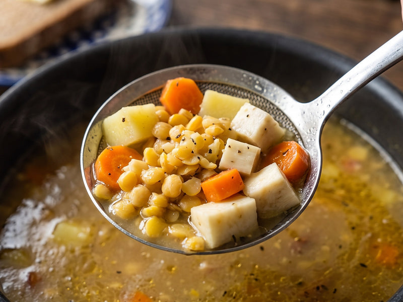 A close-up view of a metal skimmer spoon scooping up a rich mixture of split peas, diced potatoes, and root vegetables from a pot of Grochówka.