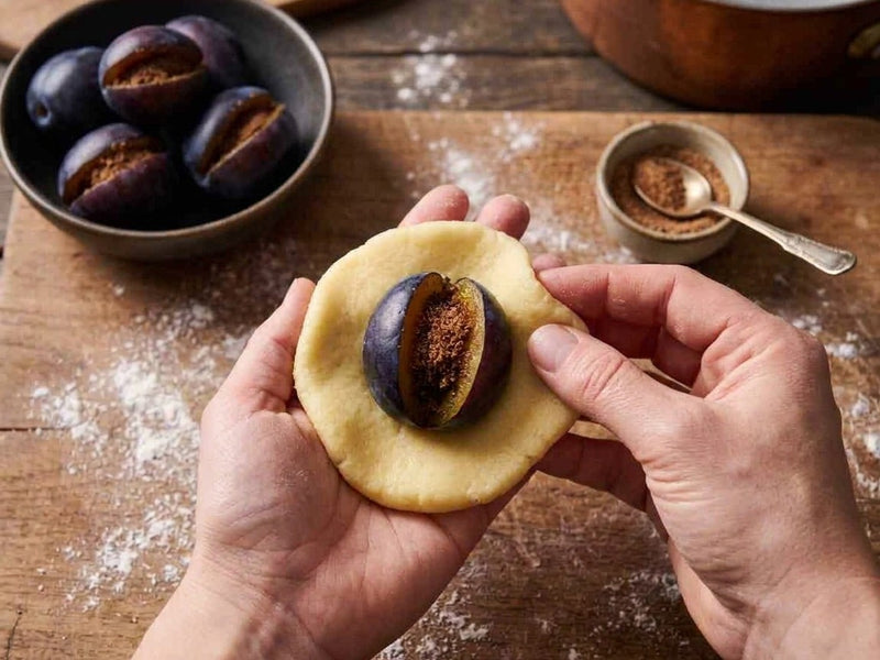 A pair of hands wrapping soft potato dough around a fresh prune plum stuffed with cinnamon and sugar.