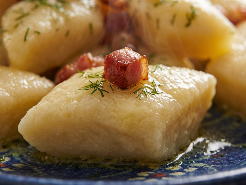 A single diamond-shaped Polish potato dumpling (Kopytko) topped with a piece of crispy bacon and fresh dill, glistening with melted butter.