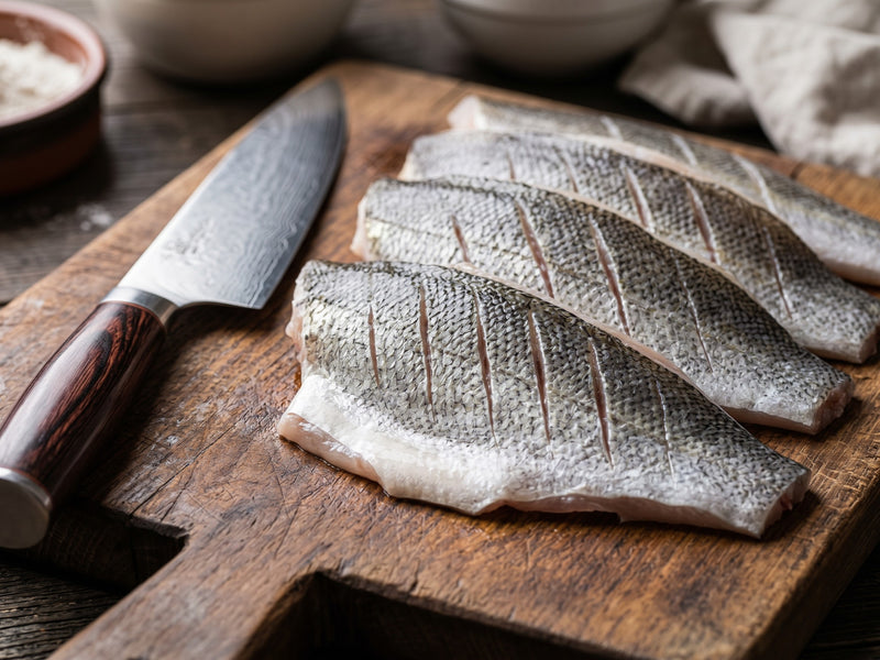 Four raw, scored zander fillets with silver skin resting on a rustic wooden chopping board next to a chef's knife and a small bowl of flour.