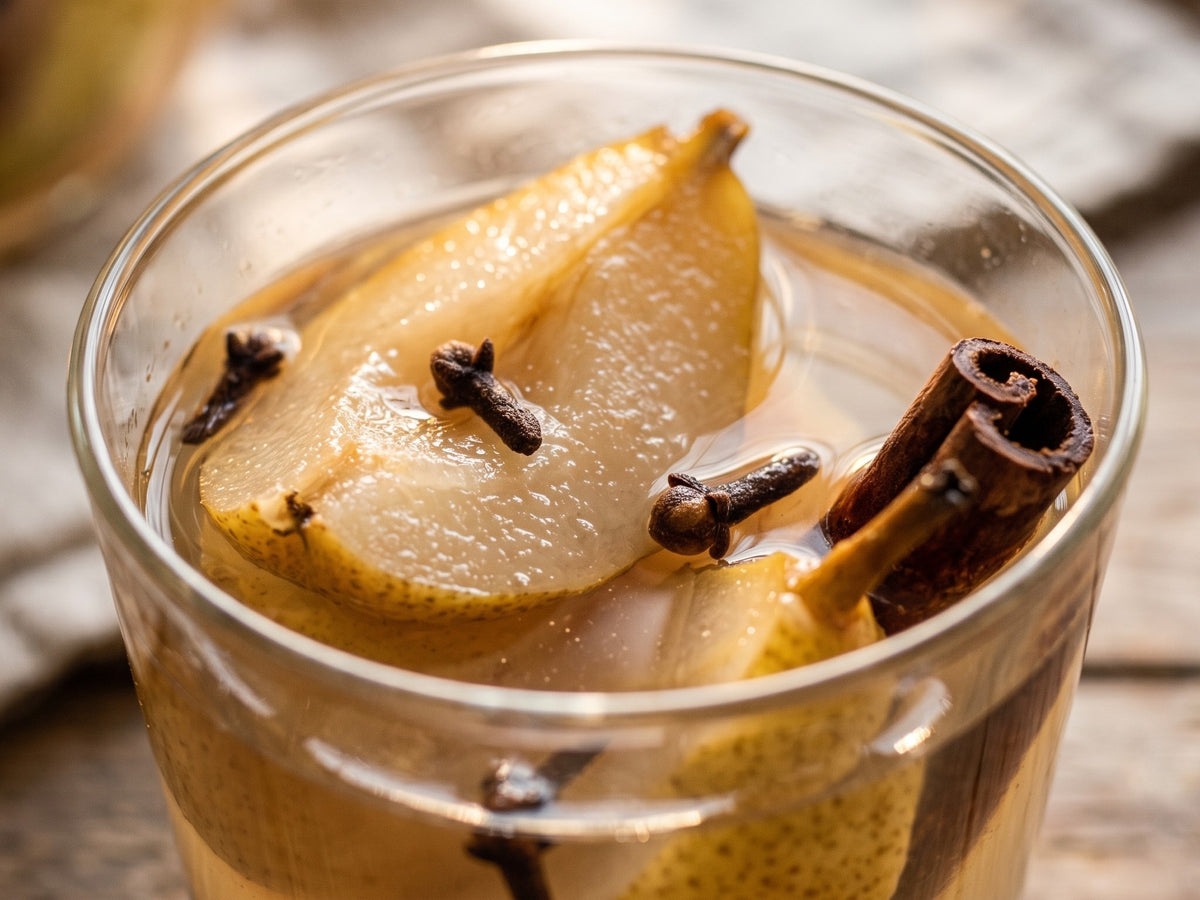 A close-up view of a glass tumbler filled with Polish pear kompot, showing steeped pear quarters, whole cloves, and a cinnamon stick.